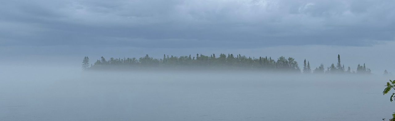 Artist's Point, wooded and dark, showing above a bank of fog on the water, seen from a gravel beach with a split-rail fence