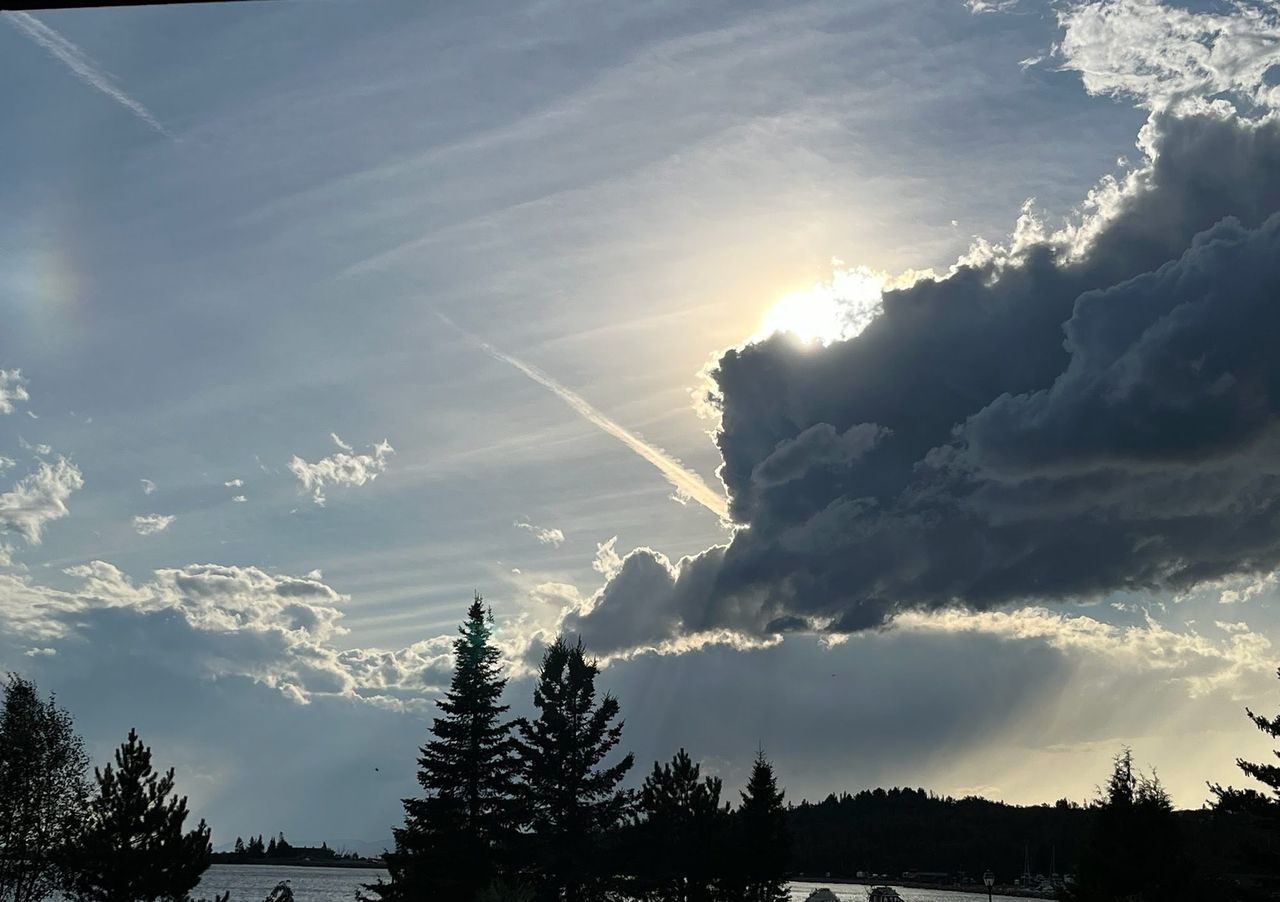 The sun behind a dark cloud, with rays falling over the harbor and boats at their moorings, seen past spruce silhouettes